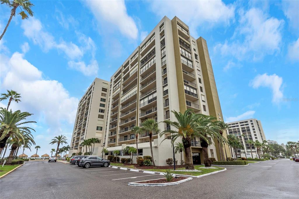 A multi-story residential building with balconies stands next to a parking lot and palm trees — 1480 GULF BOULEVARD UNIT 506, CLEARWATER BEACH, FL