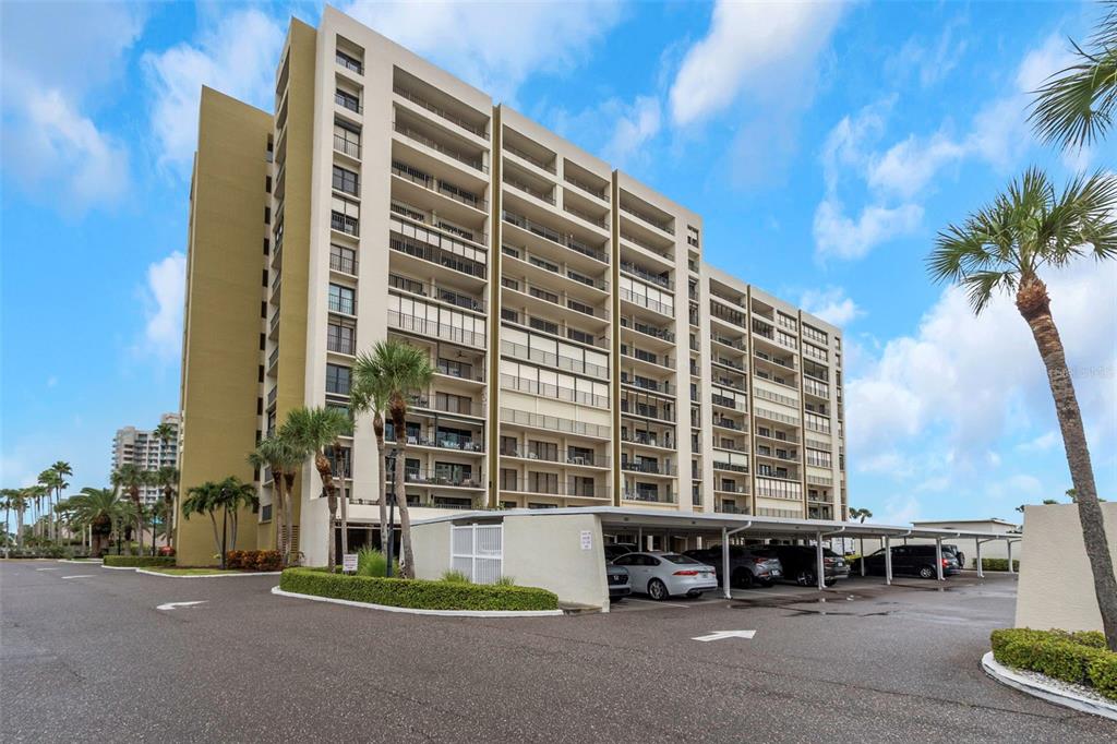 A multi-story residential building with covered parking and palm trees under a blue sky — 1480 GULF BOULEVARD UNIT 506, CLEARWATER BEACH, FL