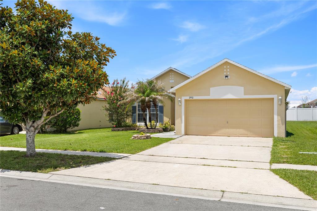 A single-story tan house with a two-car garage and a large tree in the yard — 2912 BELLE LANE, KISSIMMEE, FL