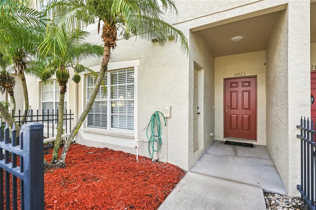 A red front door is recessed into a beige stucco exterior with a palm tree nearby — 11023 WINTER CREST DRIVE, RIVERVIEW, FL