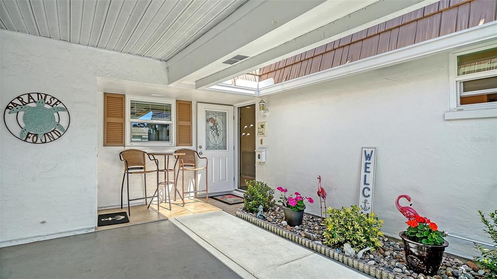 A covered patio features a small bistro set, a welcome sign, and potted plants — 2721 RIVERBLUFF PLACE UNIT V-50, SARASOTA, FL