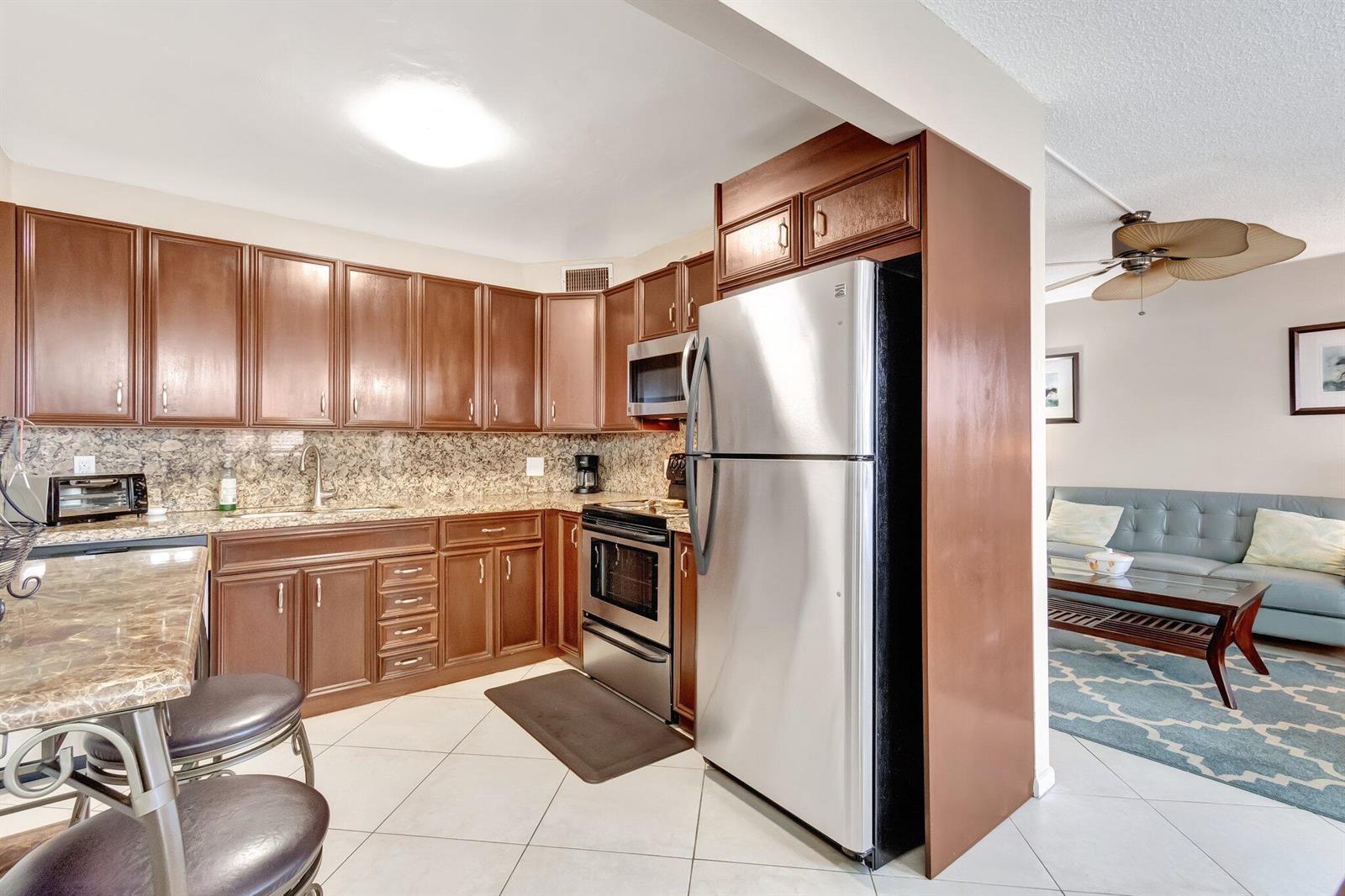 A kitchen with brown cabinets, stainless steel appliances, and granite countertops opens to a living area — 4049 Wolverton C, Boca Raton FL 33434, BOCA RATON, FL