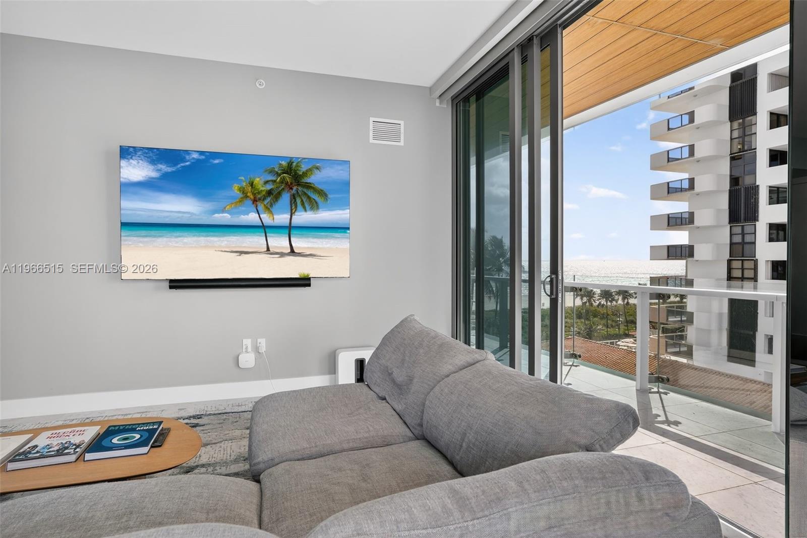 A living room features a wall-mounted television, gray sofa, and a balcony overlooking the ocean — 5775 Collins Ave # 506, Miami Beach FL 33140, MIAMI BEACH, FL