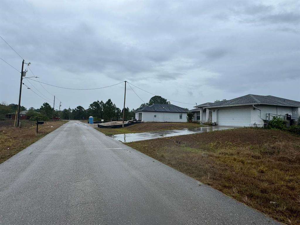 A paved road leads past a new house under construction and an existing suburban home — 1142 ECRUE STREET E, LEHIGH ACRES, FL