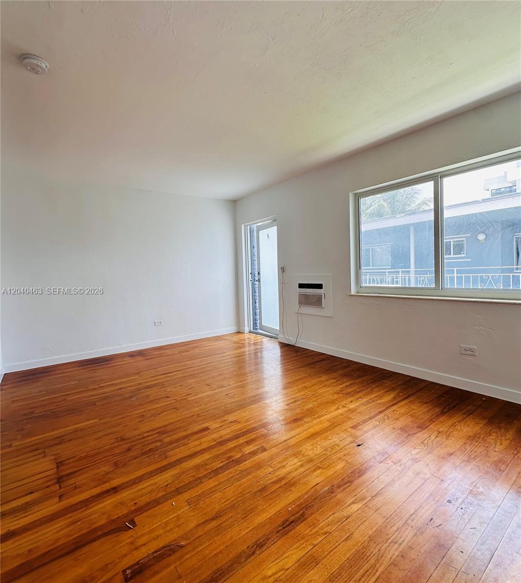 An empty room features hardwood floors, white walls, a window, and a glass-paneled door — 2045 Calais Dr # 7, Miami Beach FL 33141, MIAMI BEACH, FL