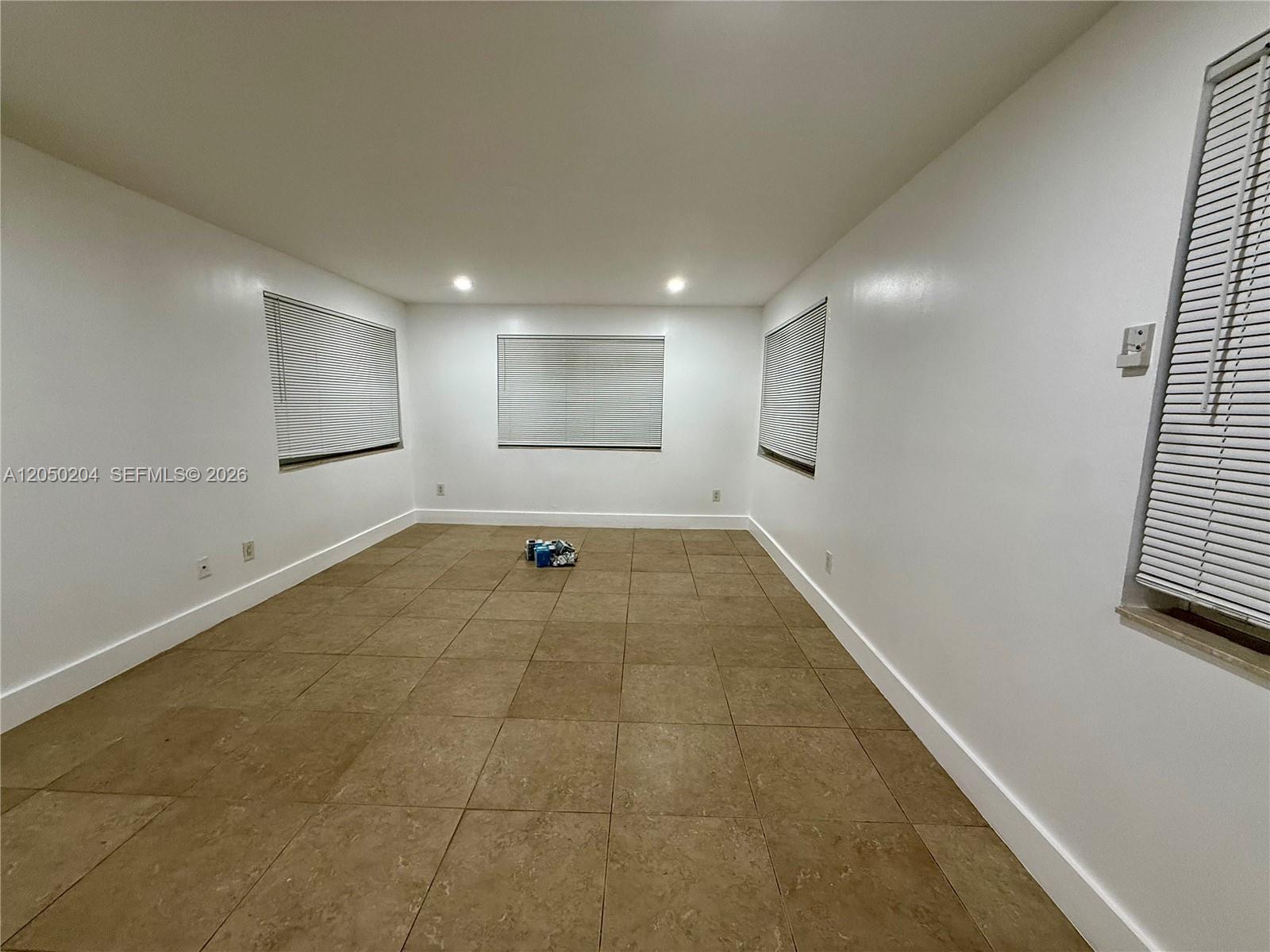 An empty room features white walls, brown tile flooring, and four windows with blinds — 427 NE 62nd St # 14, Miami FL 33138, MIAMI, FL