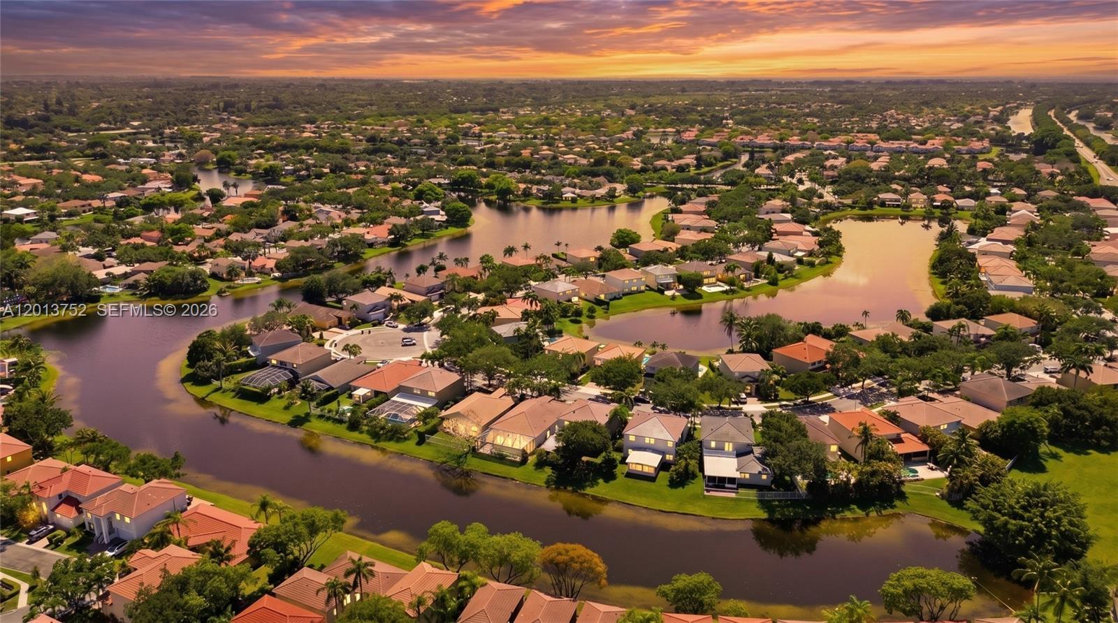 An aerial view shows a suburban neighborhood with houses surrounding a winding lake at sunset — 4019 Turquoise Trl, Weston FL 33331, WESTON, FL