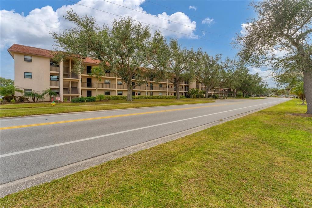 A multi-story apartment building with balconies stands beside a paved road and grassy lawn — 104 CAPRI ISLES BOULEVARD UNIT 112, VENICE, FL