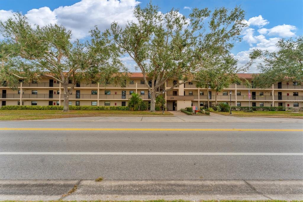 A three-story apartment building with a tan exterior sits behind a road and trees — 104 CAPRI ISLES BOULEVARD UNIT 112, VENICE, FL