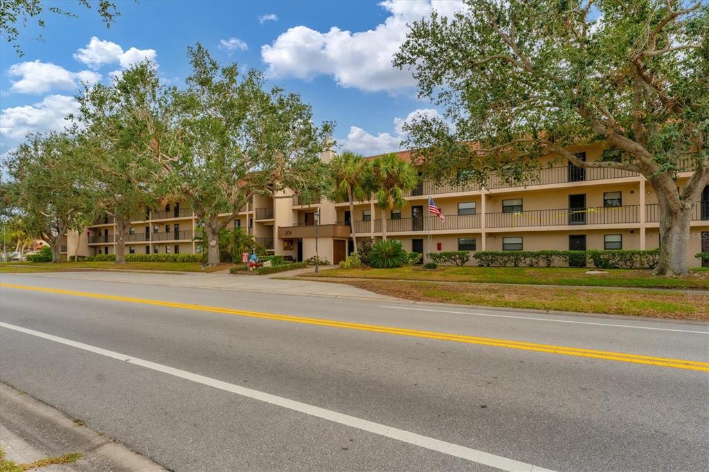 A multi-story apartment building with balconies stands behind a road and large trees — 104 CAPRI ISLES BOULEVARD UNIT 112, VENICE, FL