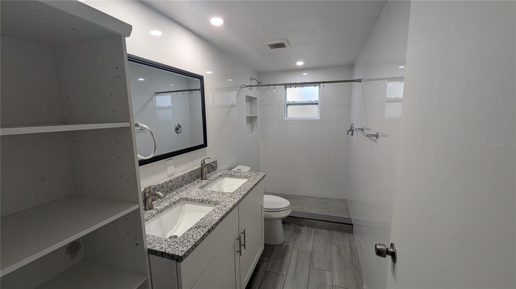 A modern bathroom features a double vanity with granite countertops, a walk-in shower, and shelving — 5723 9TH STREET CIRCLE E, BRADENTON, FL