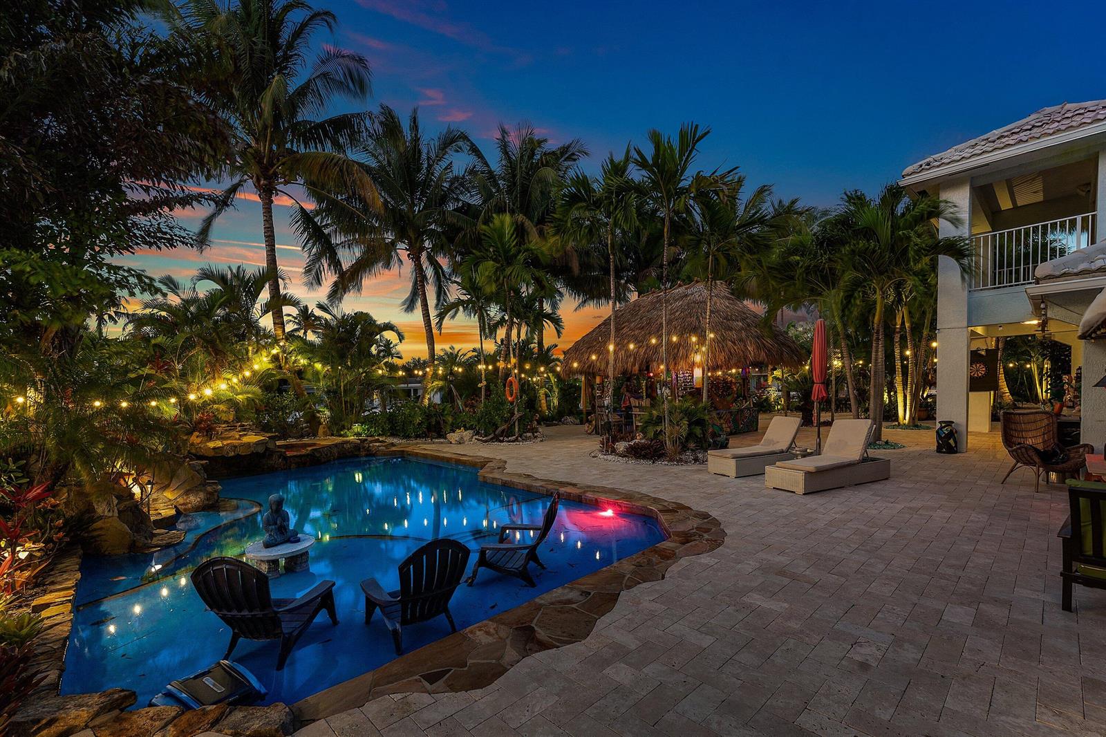 A backyard pool area features a tiki hut, lounge chairs, and string lights at dusk — 2311 NE 47th Street, Lighthouse Point FL 33064, LIGHTHOUSE POINT, FL