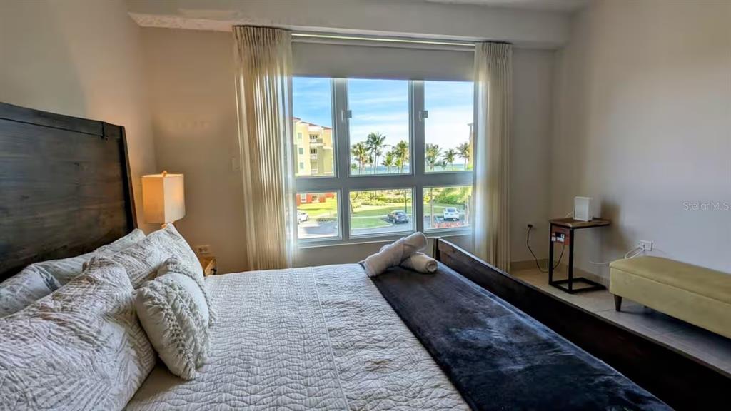 A bedroom features a wooden bed frame, white bedding, and a large window overlooking palm trees — 100 OCEAN DRIVE UNIT 131, HUMACAO, PR