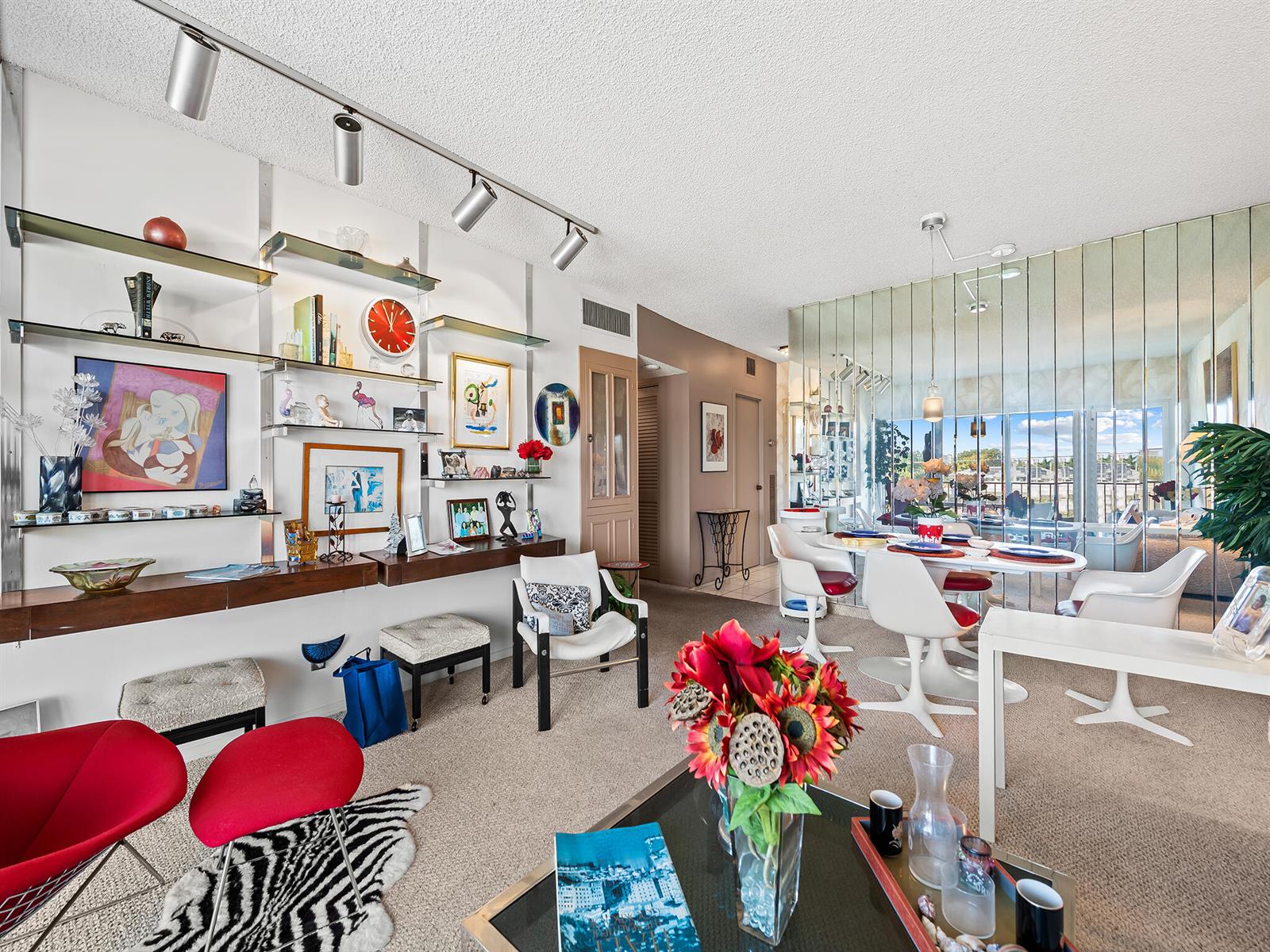 A living and dining room features a wall-mounted shelving unit, mirrored wall, and carpeted floors — 2500 E Las Olas Boulevard # 404, Fort Lauderdale FL 33301, FORT LAUDERDALE, FL