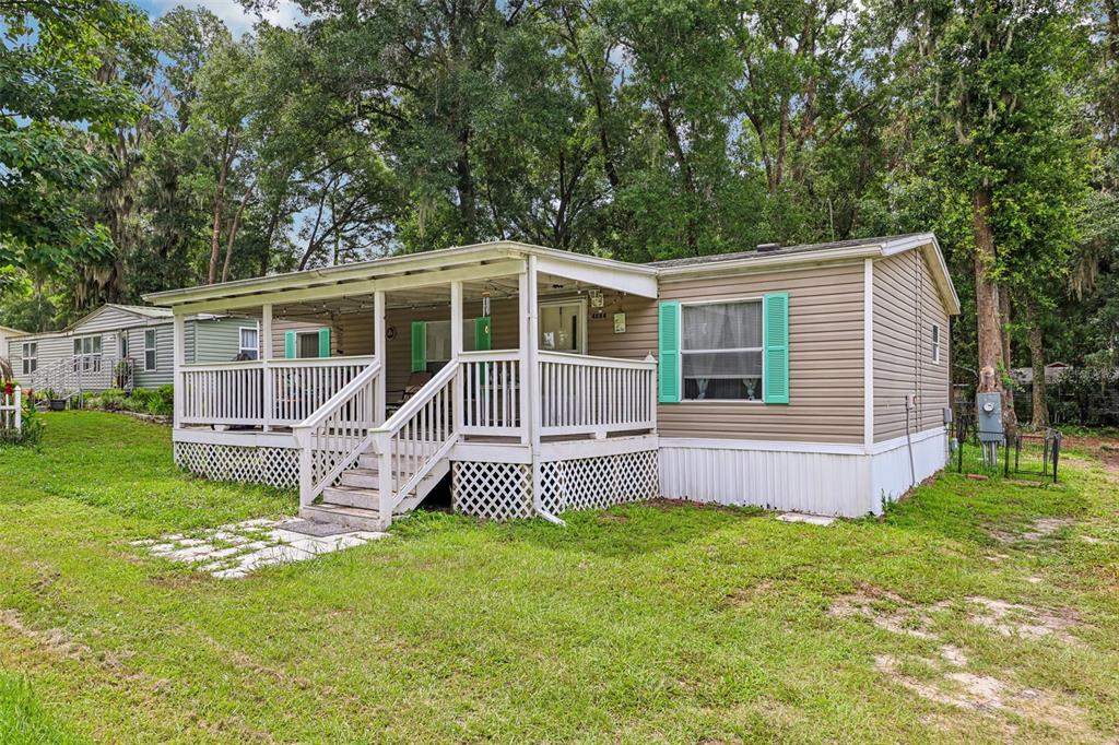 A tan mobile home with a white covered porch and teal shutters sits on a lawn — 4884 SE 146TH LANE, SUMMERFIELD, FL