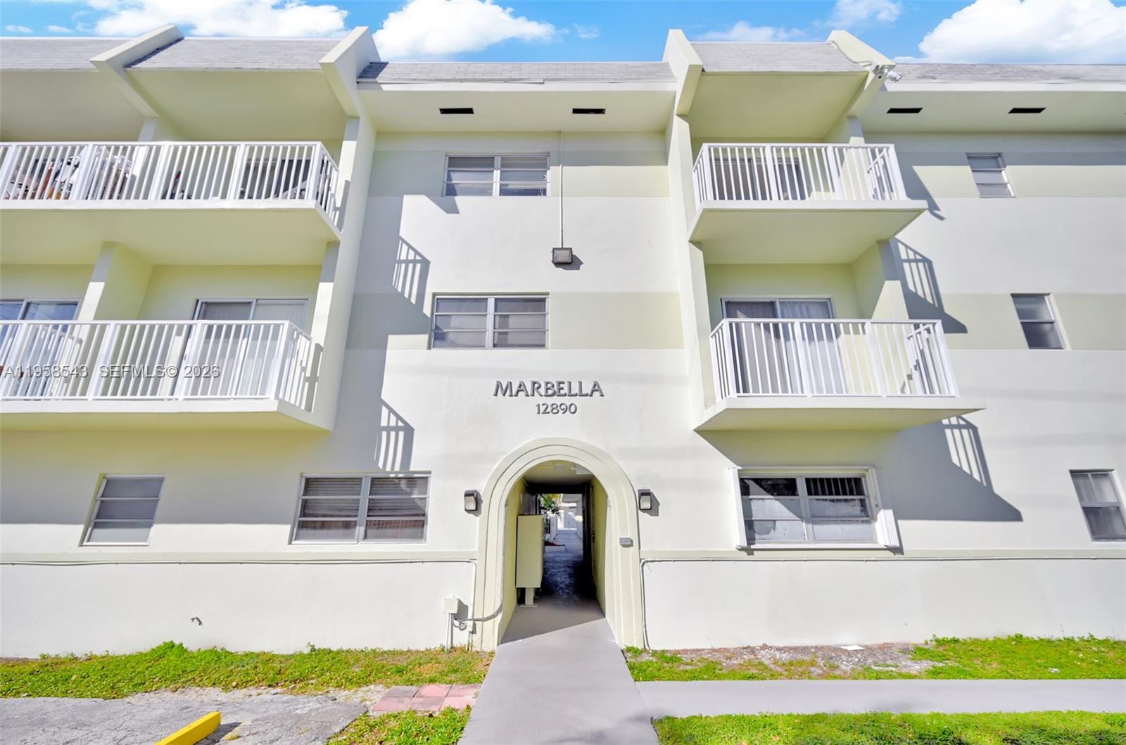 The exterior of a three-story apartment building features white balconies and a central arched entryway — 12890 NE 8th Ave # 208, North Miami FL 33161, NORTH MIAMI, FL