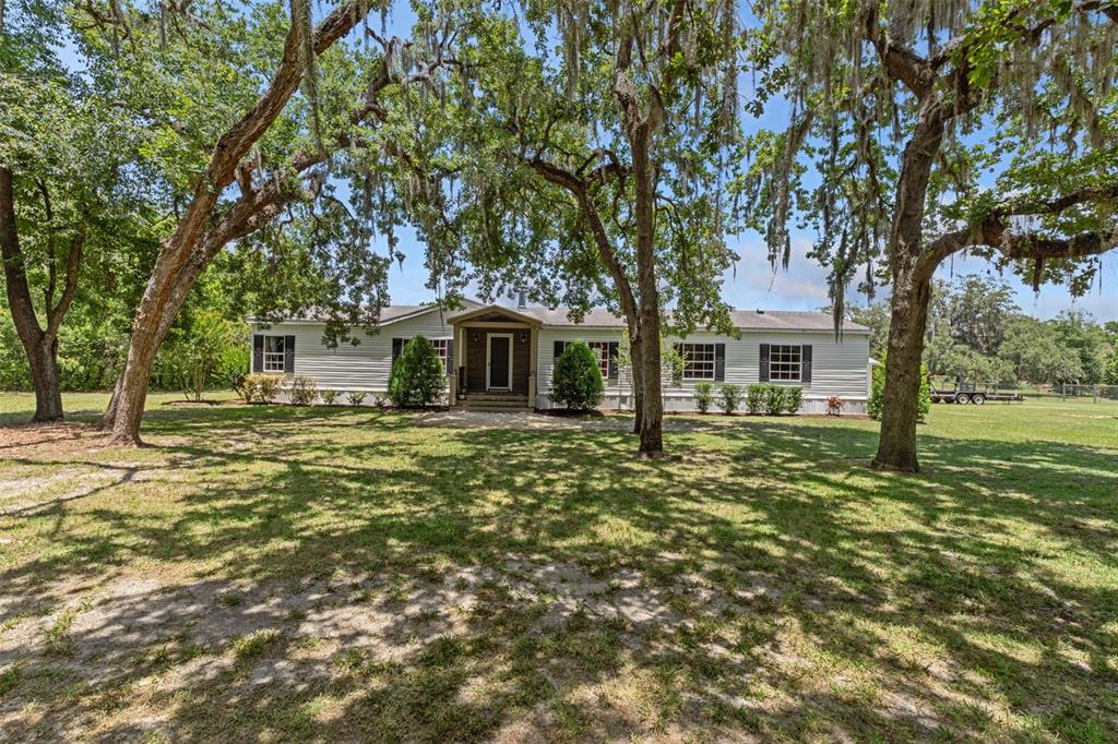 A single-story manufactured home with light siding sits on a grassy lot under large trees — 26255 LAMBETH ROAD, BROOKSVILLE, FL