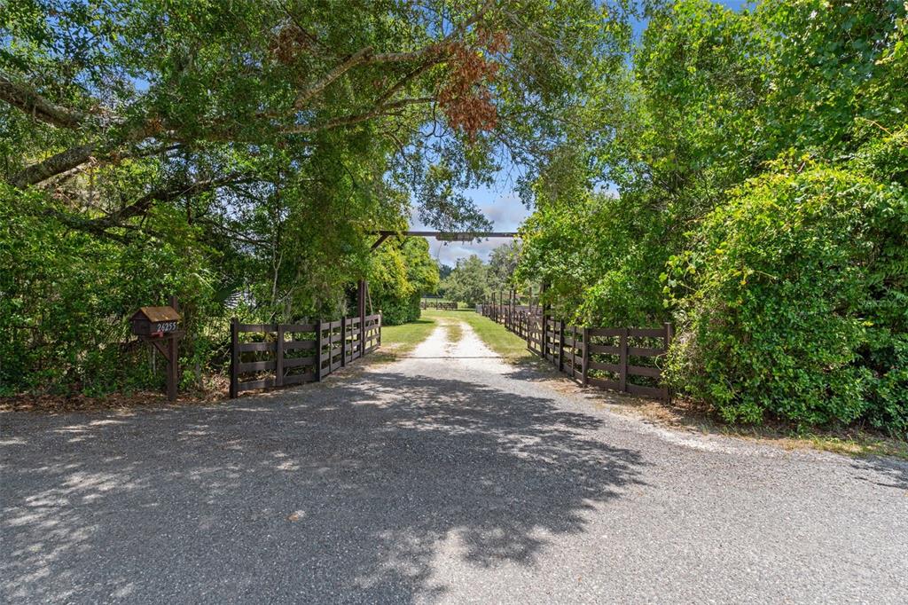 A gravel driveway leads through a wooden gate entrance flanked by trees and fences — 26255 LAMBETH ROAD, BROOKSVILLE, FL