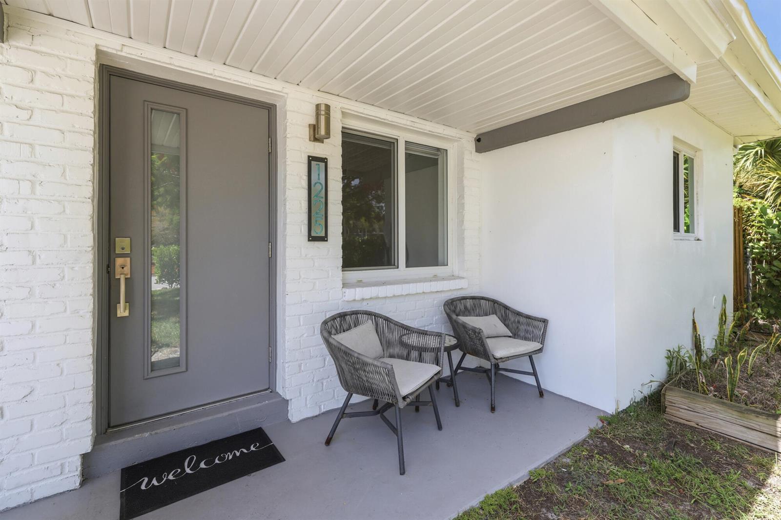 A covered front porch features a gray door, two wicker chairs, and white brick walls — 1225 15th Avenue N, FL 33460, FL