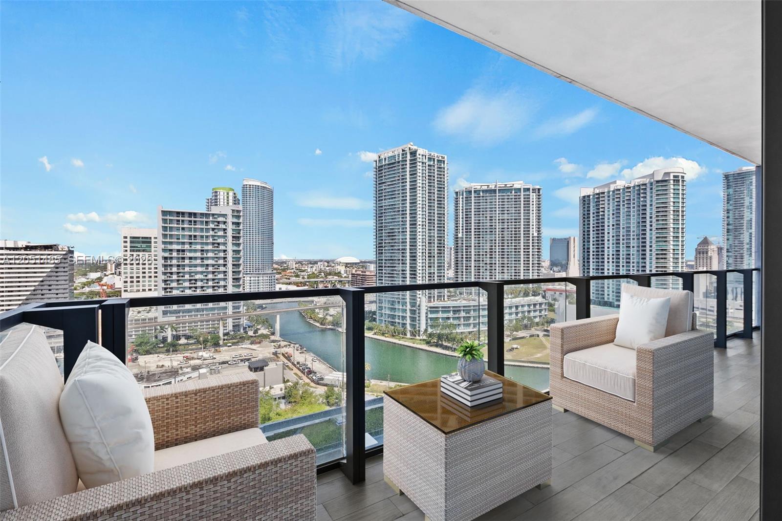 A balcony with wicker chairs and a table overlooks a river and city skyline — 68 SE 6th St # 1706, Miami FL 33131, MIAMI, FL