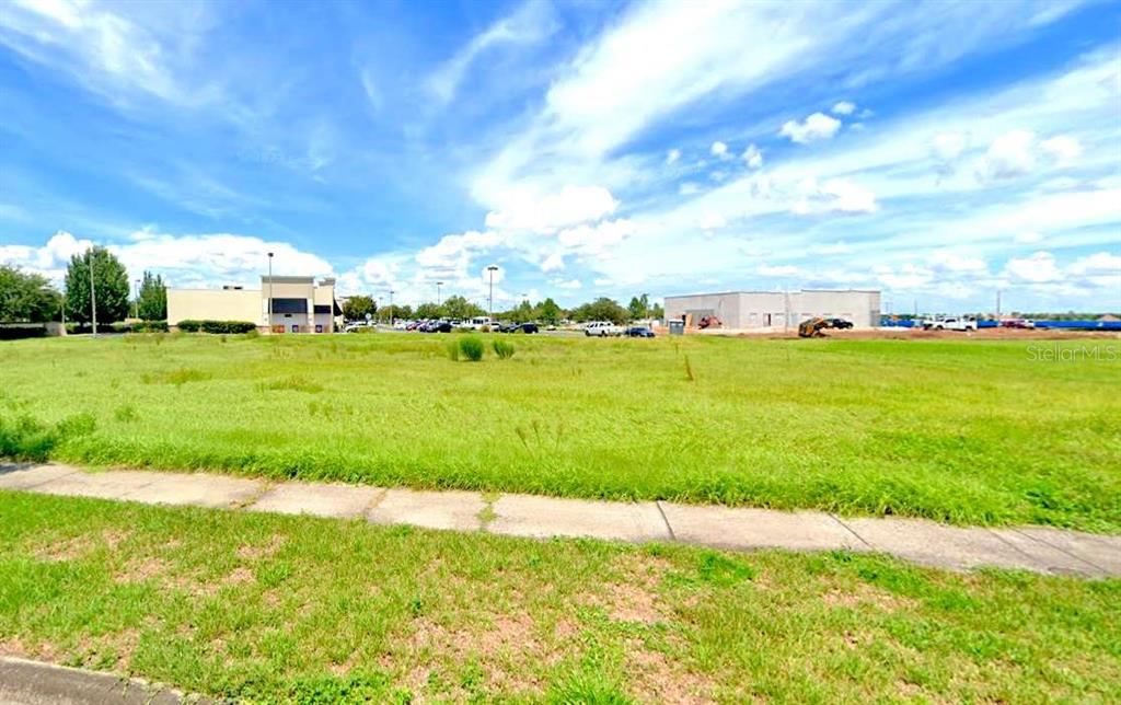 A large, grassy vacant lot sits in front of commercial buildings under a blue sky — 0 BALL PARK ROAD, KISSIMMEE, FL