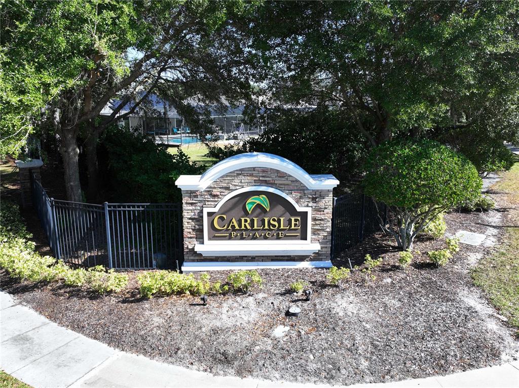 An exterior stone monument sign identifies the Carlisle Place community near a black metal fence — 13822 BASSINGTHORPE DRIVE, SPRING HILL, FL
