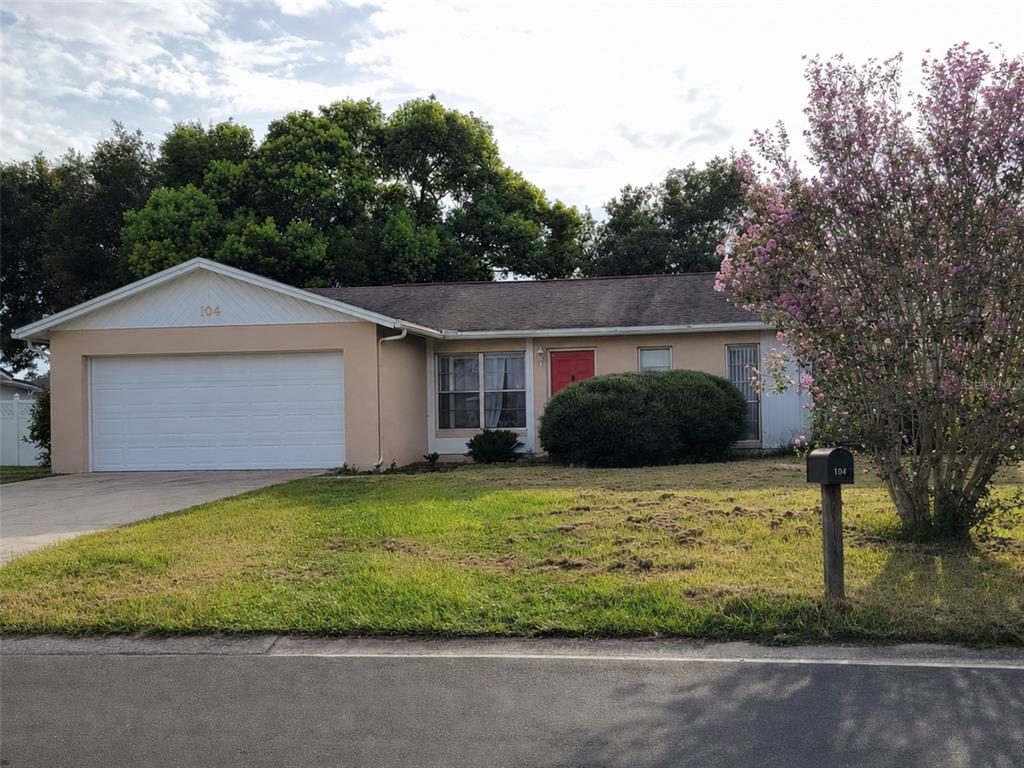A single-story house with a beige exterior, white garage door, red front door, and flowering tree — 104 MORELIA LANE, KISSIMMEE, FL