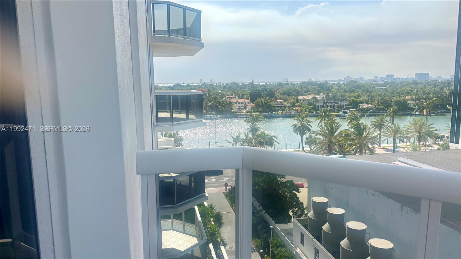 A high-angle view from a balcony overlooks a waterway, palm trees, and distant city buildings — 4779 Collins Ave # 808, Miami Beach FL 33140, MIAMI BEACH, FL