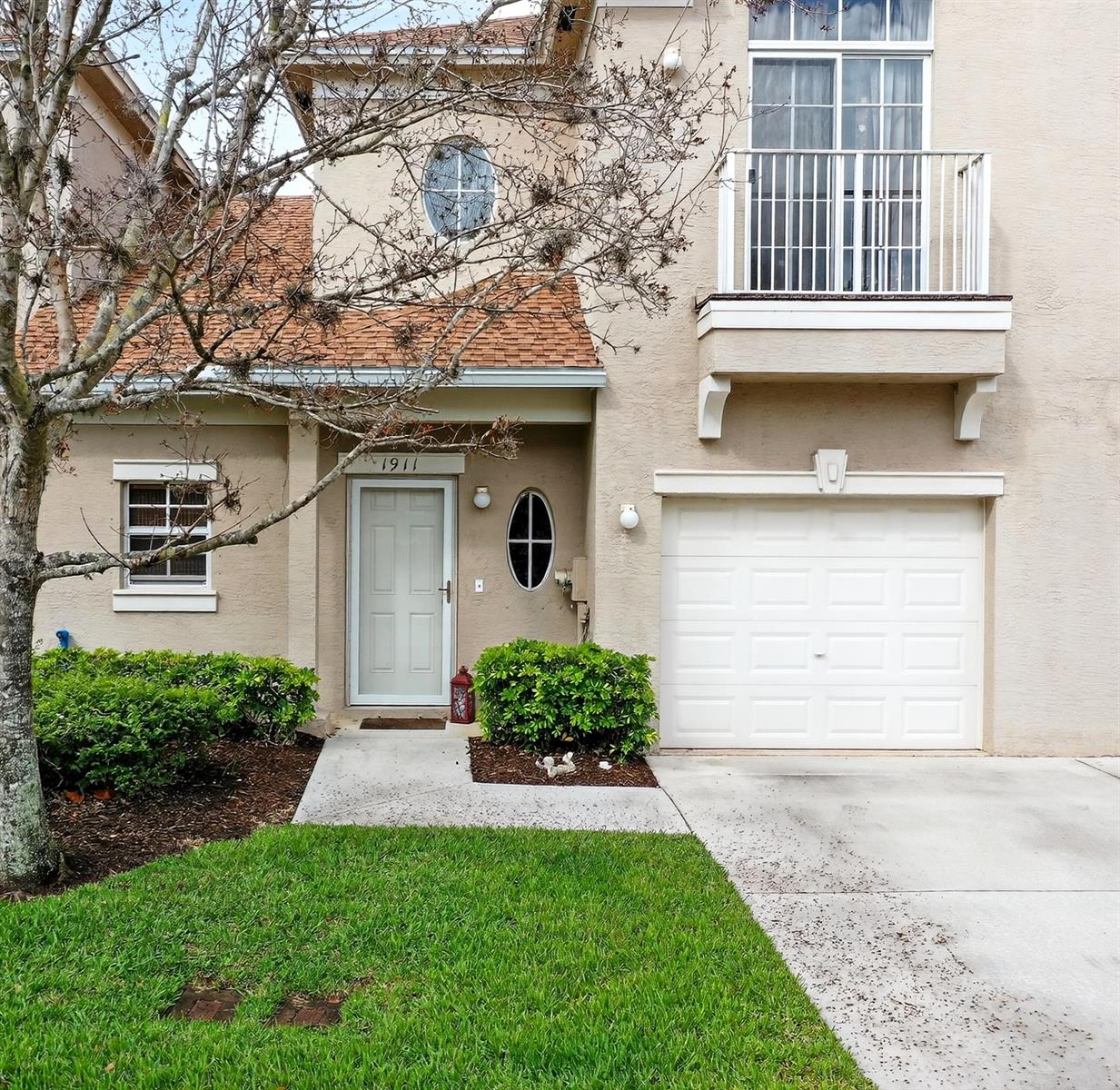 A two-story tan stucco townhouse features a white garage door, balcony, and front entrance — 1911 77th Drive, Vero Beach FL 32966, VERO BEACH, FL