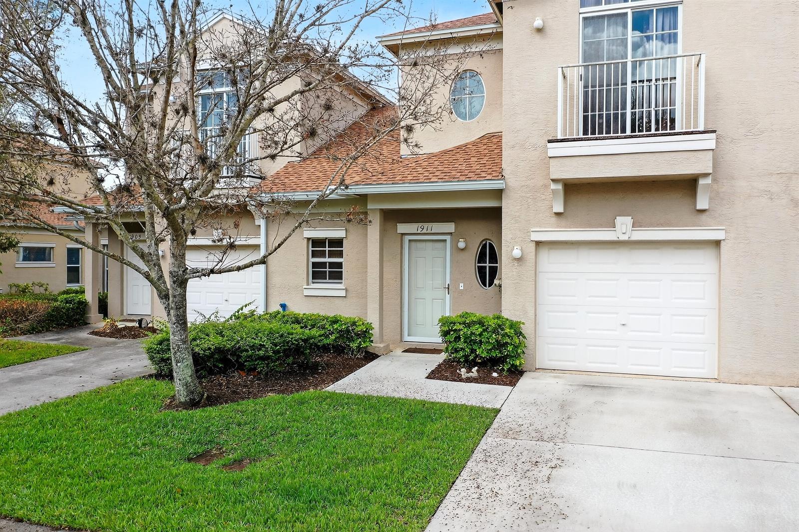 The exterior of a two-story beige townhouse features a garage, a balcony, and landscaping — 1911 77th Drive, Vero Beach FL 32966, VERO BEACH, FL