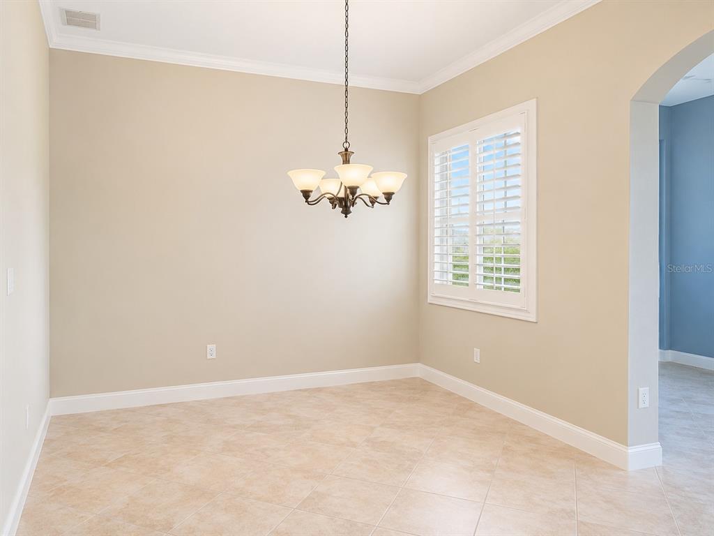 An empty dining room features beige walls, tile flooring, a chandelier, and plantation shutters — 8361 MIRAMAR WAY, LAKEWOOD RANCH, FL