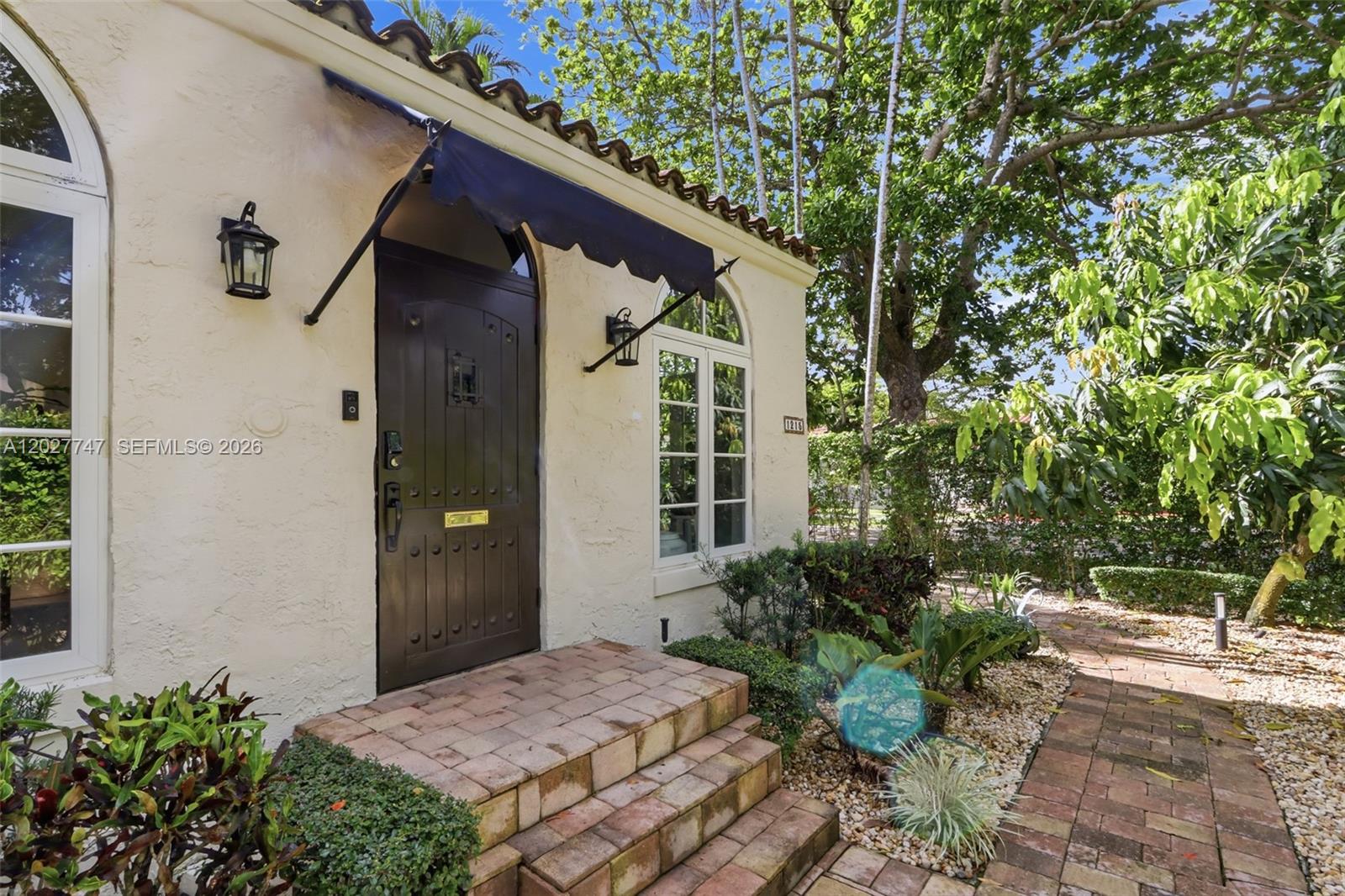 The exterior of a stucco house features a dark arched door, brick steps, and awning — 1215 Madrid St, Coral Gables FL 33134, CORAL GABLES, FL