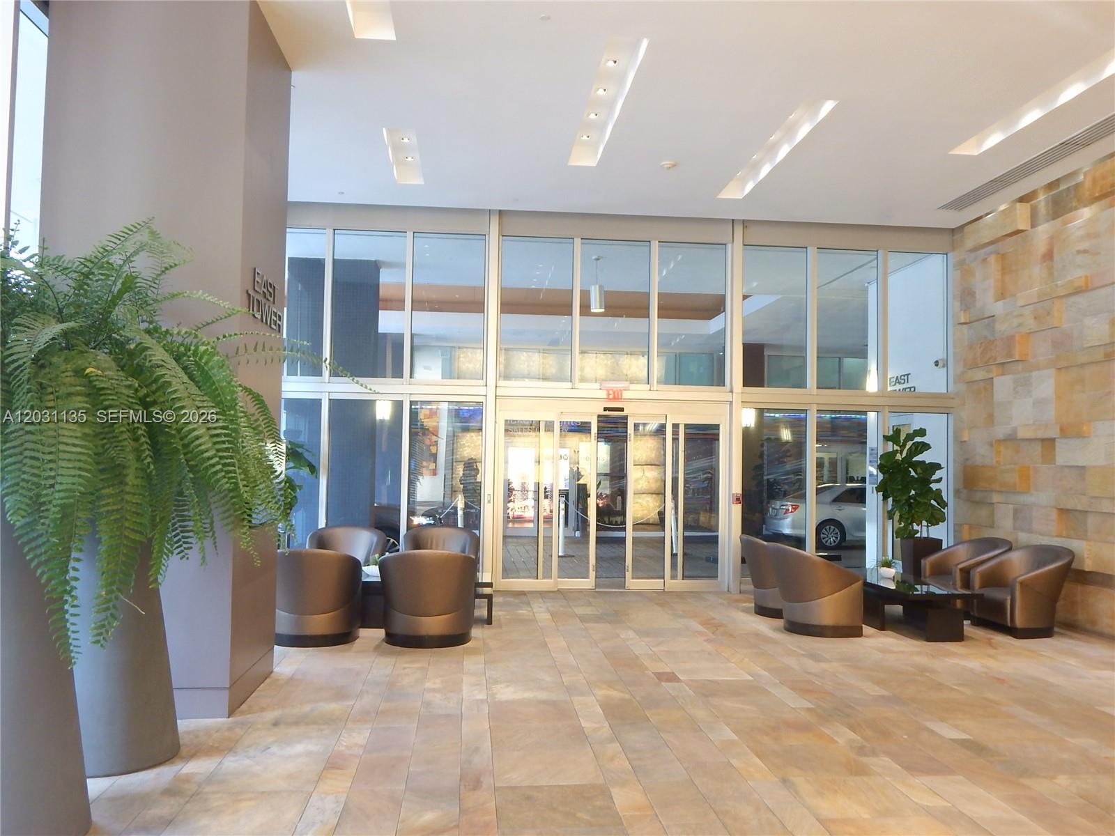 A modern building lobby features stone tile flooring, glass entrance doors, and seating areas — 500 Brickell Ave # 1808, Miami FL 33131, MIAMI, FL