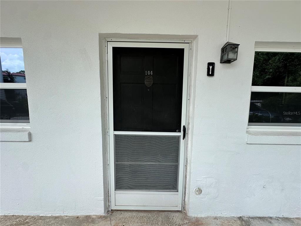 A white exterior wall features a black front door with a screen door and windows — 100 EL CAMINO DR UNIT 104, WINTER HAVEN, FL