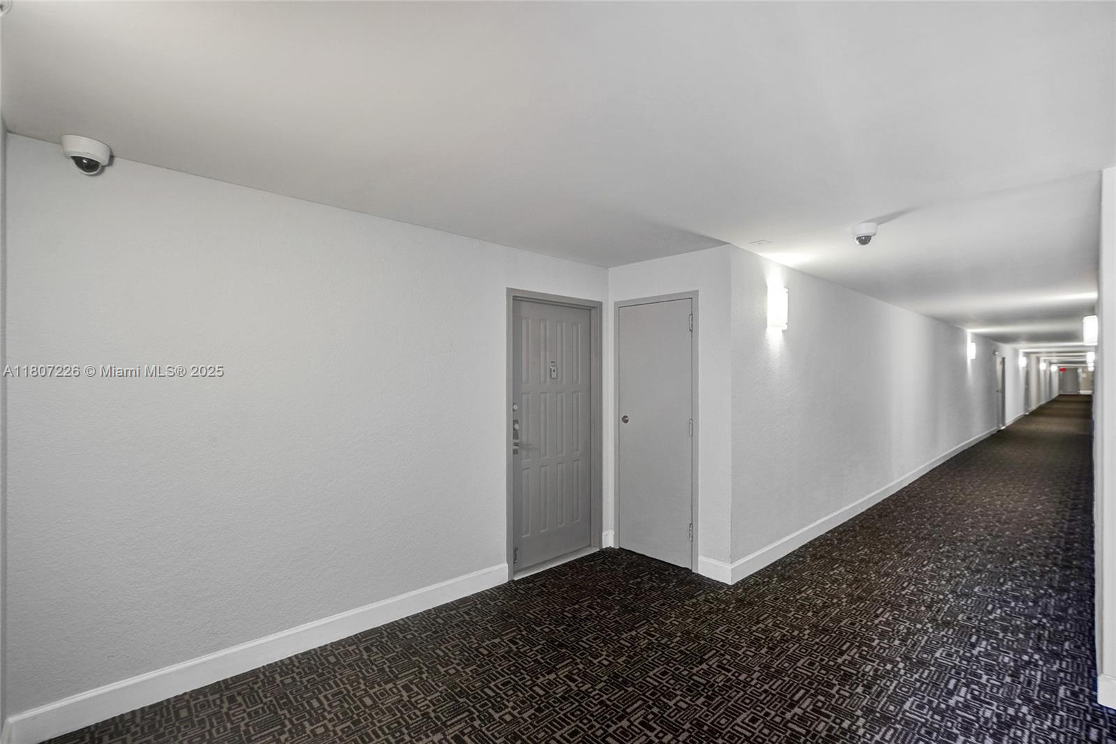 A long, carpeted apartment building hallway features white walls, two doors, and security cameras — 200 Leslie Dr # 1031, Hallandale Beach FL 33009, HALLANDALE BEACH, FL