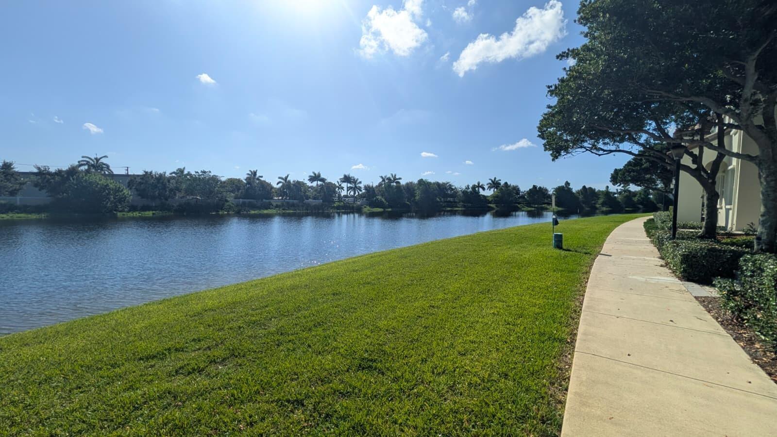 A concrete walkway runs alongside a grassy lawn bordering a calm lake under a sunny sky — 4153 Cascada Circle, Cooper City FL 33024, COOPER CITY, FL