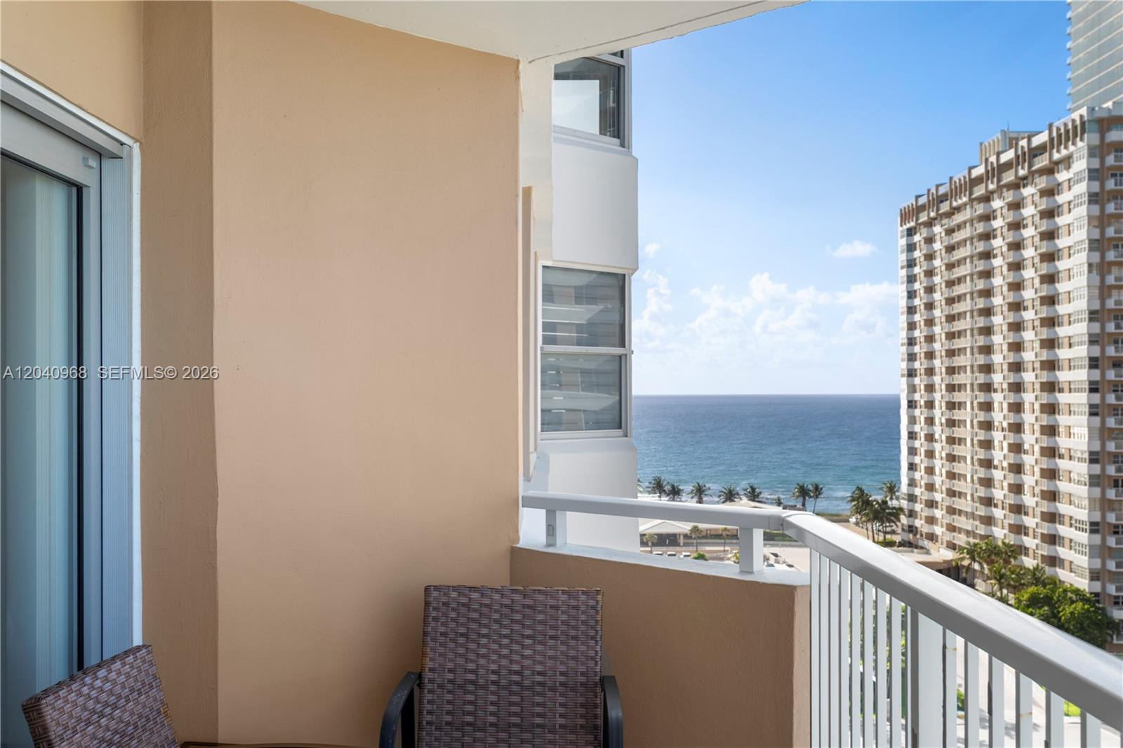 A balcony with wicker chairs overlooks the ocean and a neighboring high-rise building — 1965 S Ocean Dr # 11E, Hallandale Beach FL 33009, HALLANDALE BEACH, FL