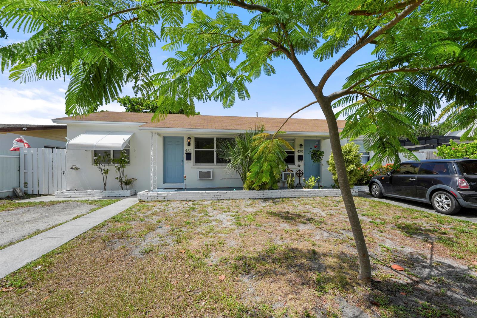 A single-story duplex with white walls, blue doors, and a brown roof sits behind trees — 429 Lytle Street, West Palm Beach FL 33405, WEST PALM BEACH, FL
