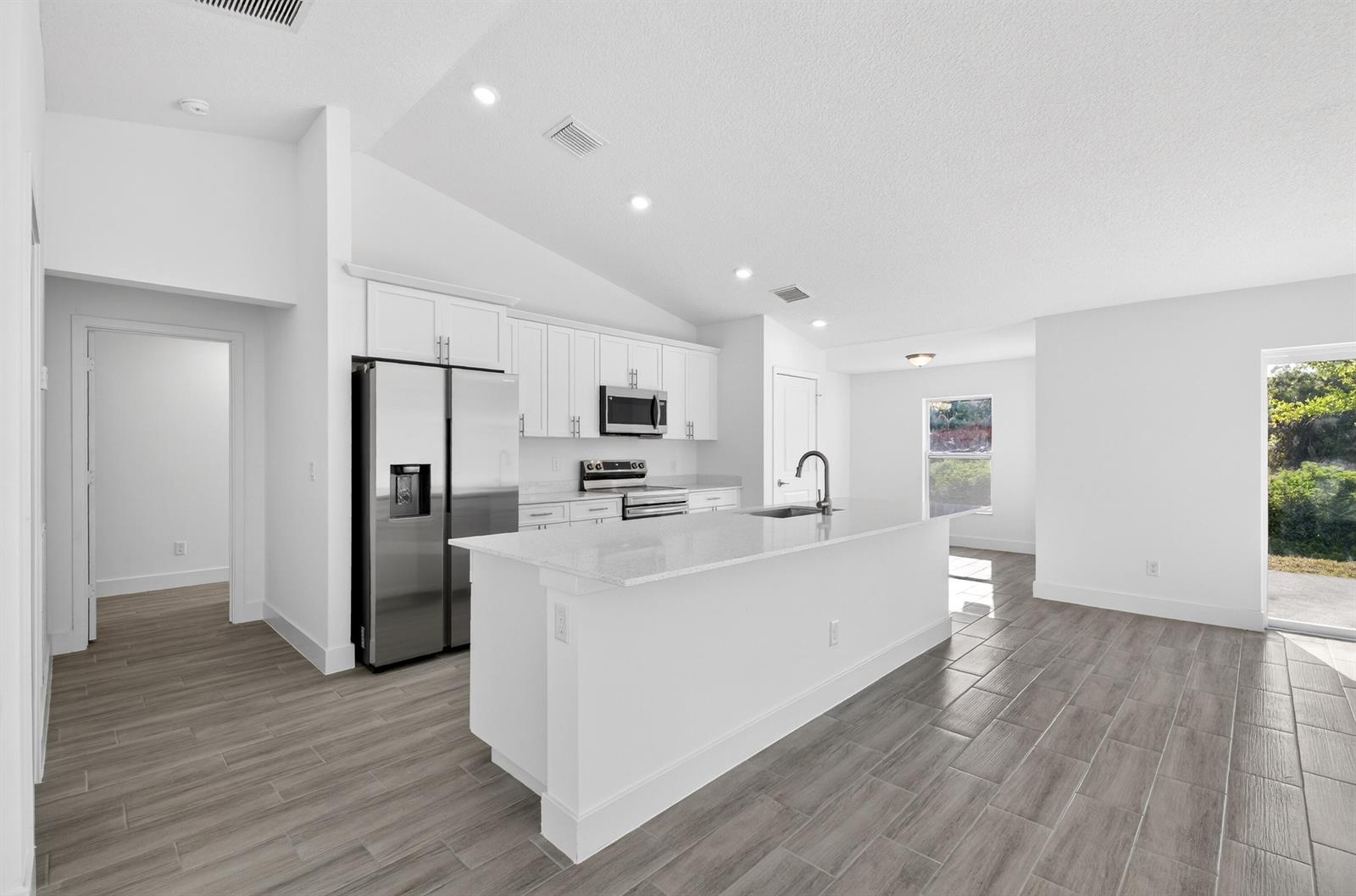 A modern kitchen features white cabinetry, stainless steel appliances, a quartz island, and wood-look flooring — 7785 105th Avenue, Vero Beach FL 32967, VERO BEACH, FL