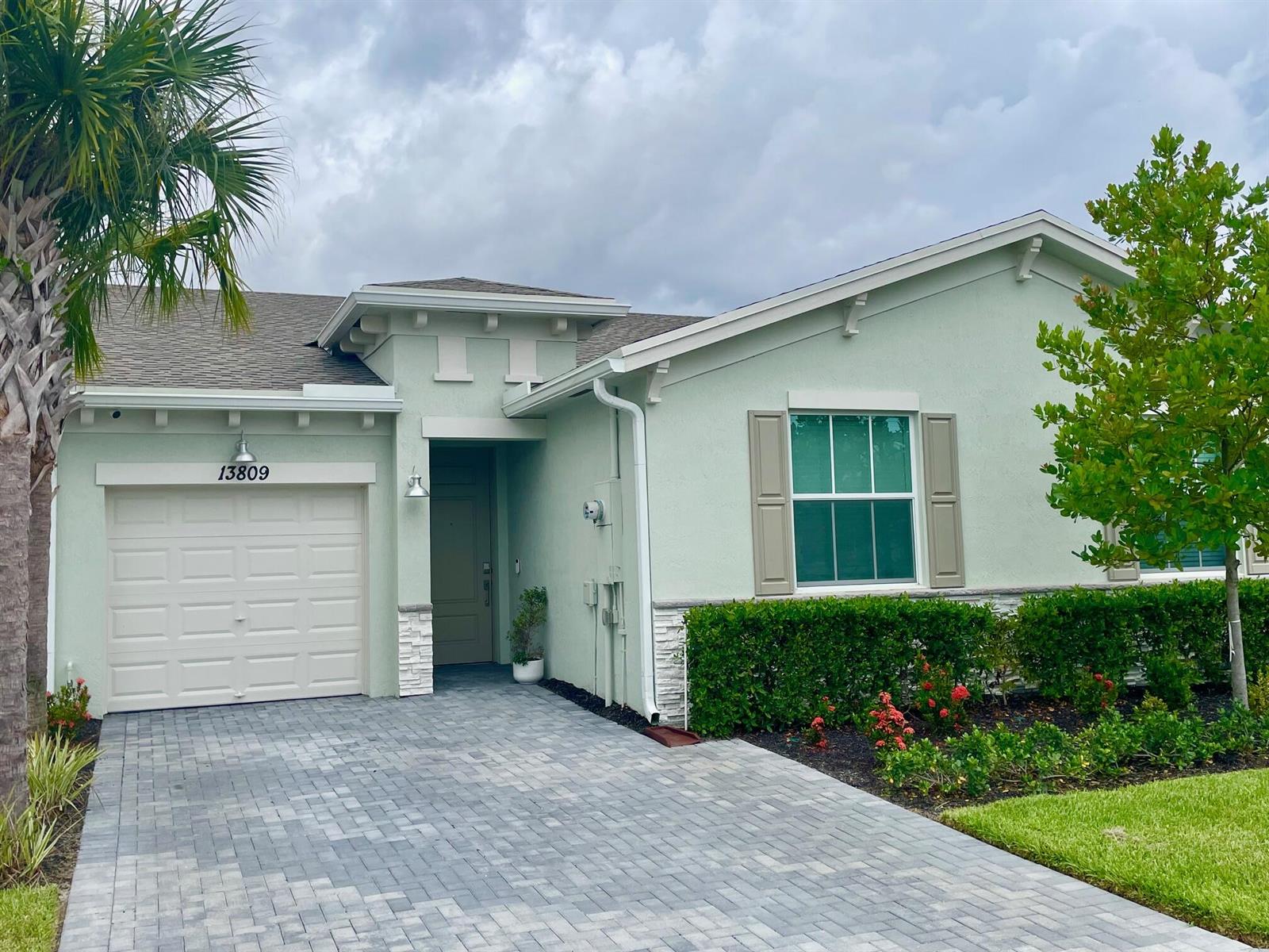 The exterior of a single-story residential building features a paved driveway, landscaping, and garage doors — 13809 Hero Path, Delray Beach FL 33484, DELRAY BEACH, FL