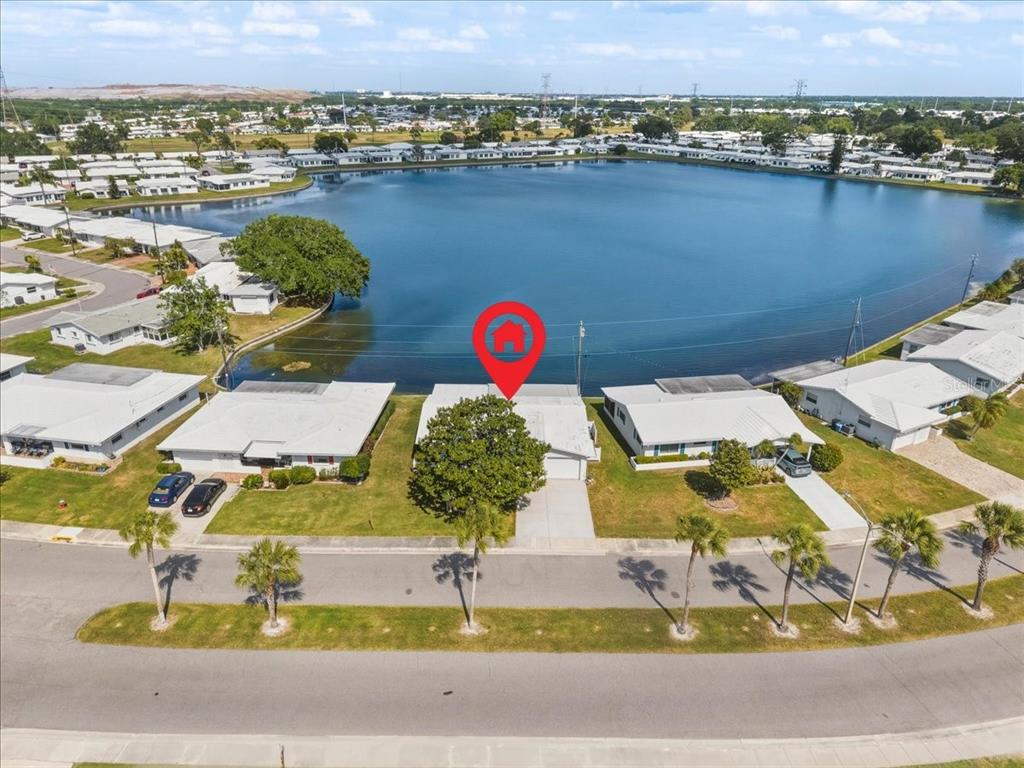 An aerial view shows a single-story house with a white roof located beside a lake — 9875 MAINLANDS BOULEVARD W, PINELLAS PARK, FL