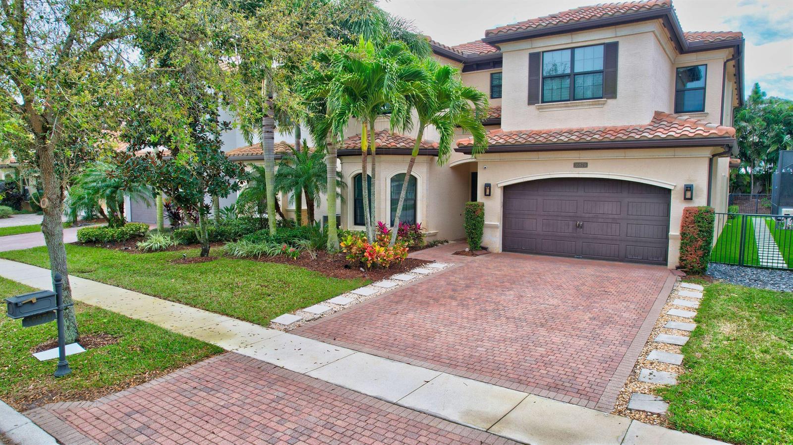 A two-story stucco house features a brick driveway, a brown garage door, and palm trees — 16879 Bridge Crossing Circle, Delray Beach FL 33446, DELRAY BEACH, FL