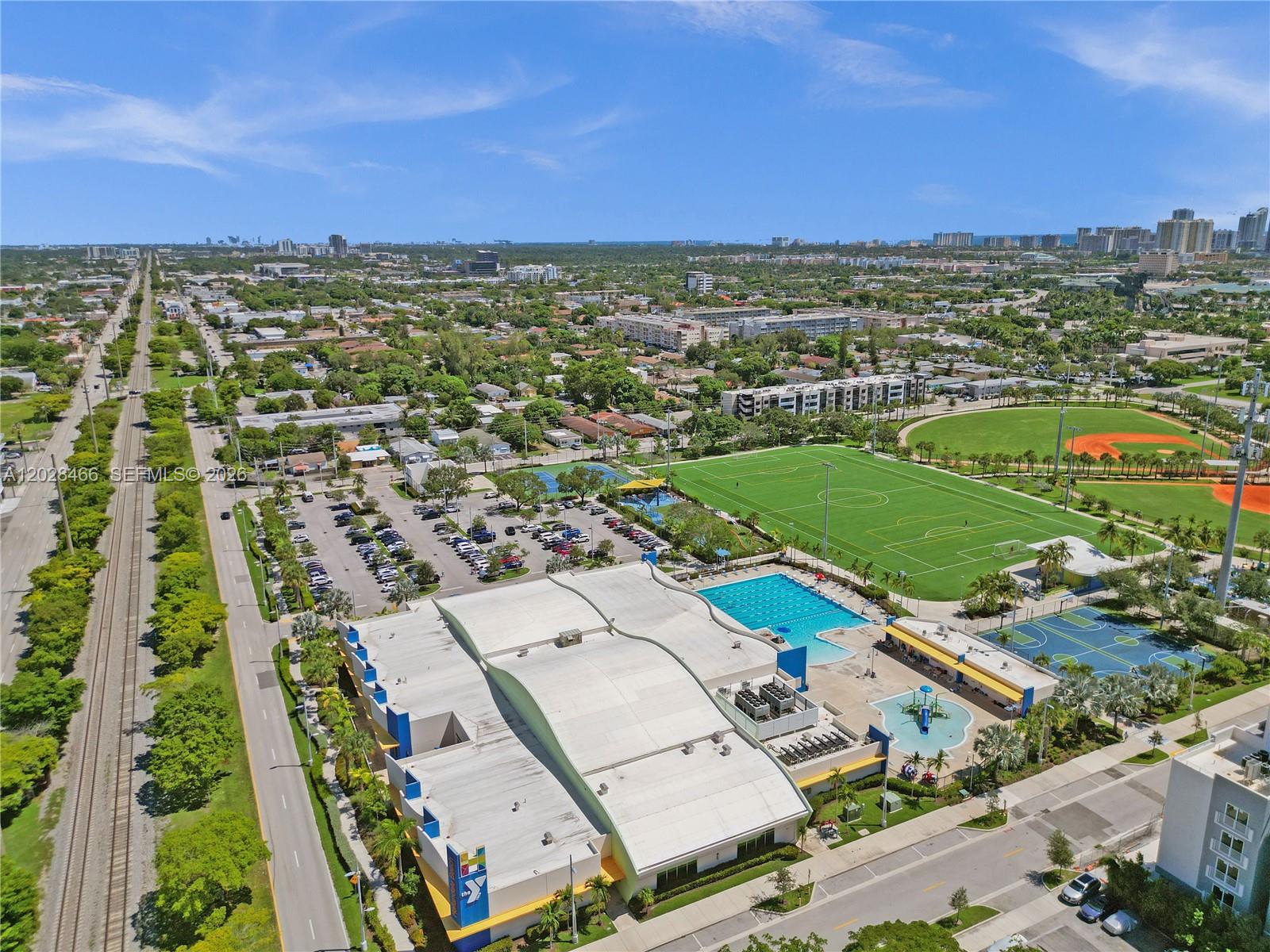 An aerial view shows a YMCA facility with a swimming pool, sports fields, and parking — 114 SE 8th St # 105, Hallandale Beach FL 33009, HALLANDALE BEACH, FL