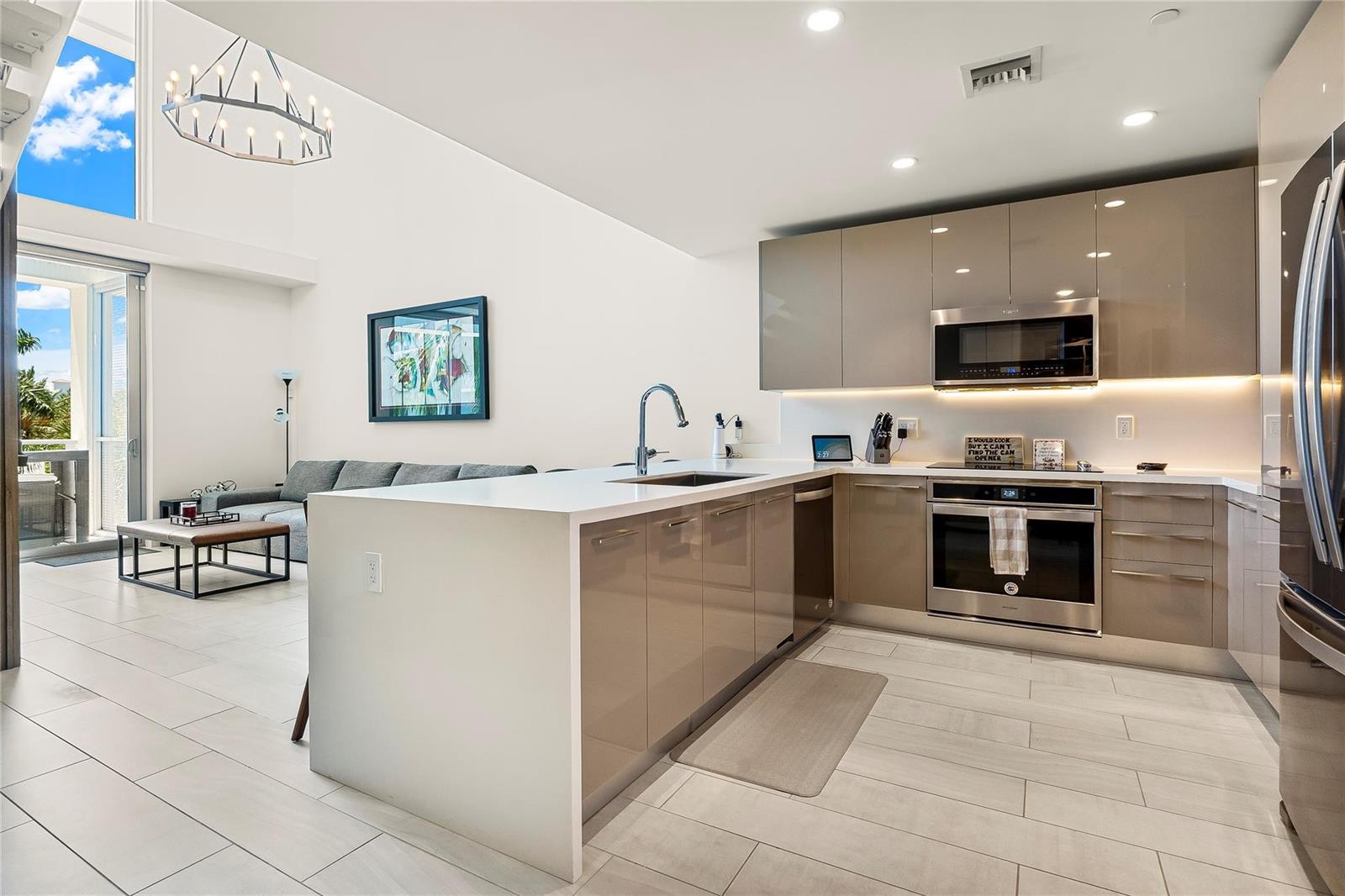A modern kitchen with glossy taupe cabinets and white countertops opens to a living area — 2000 Metropica Way # 216, Sunrise FL 33323, SUNRISE, FL