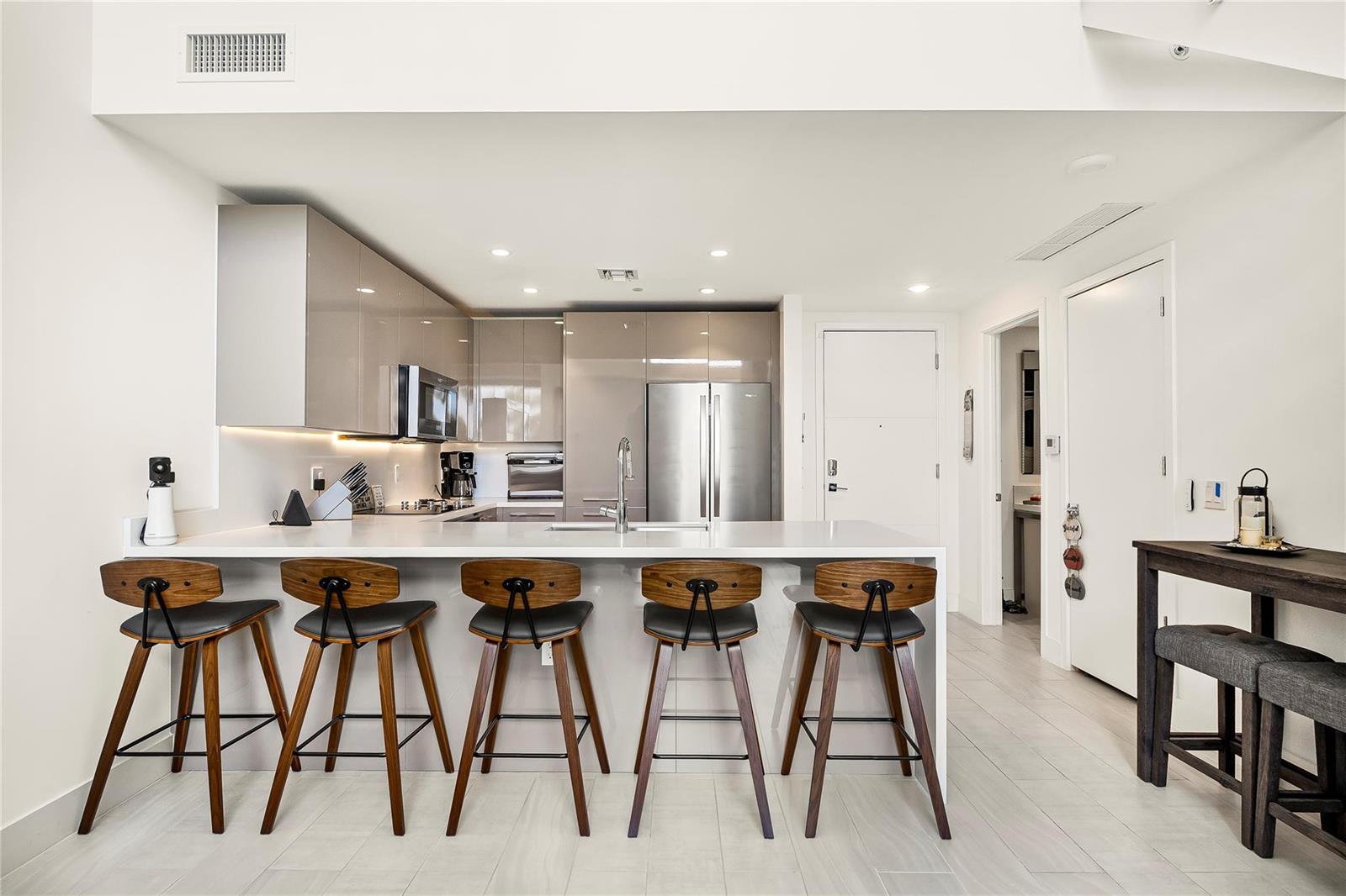 A modern kitchen features glossy taupe cabinets, stainless steel appliances, and five wooden bar stools — 2000 Metropica Way # 216, Sunrise FL 33323, SUNRISE, FL