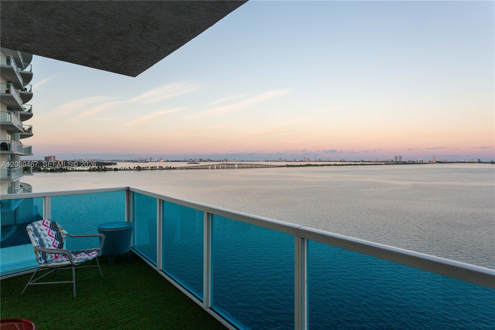 A high-rise balcony with blue glass railings overlooks a bay and a distant bridge — 700 NE 25th St # 1003, Miami FL 33137, MIAMI, FL