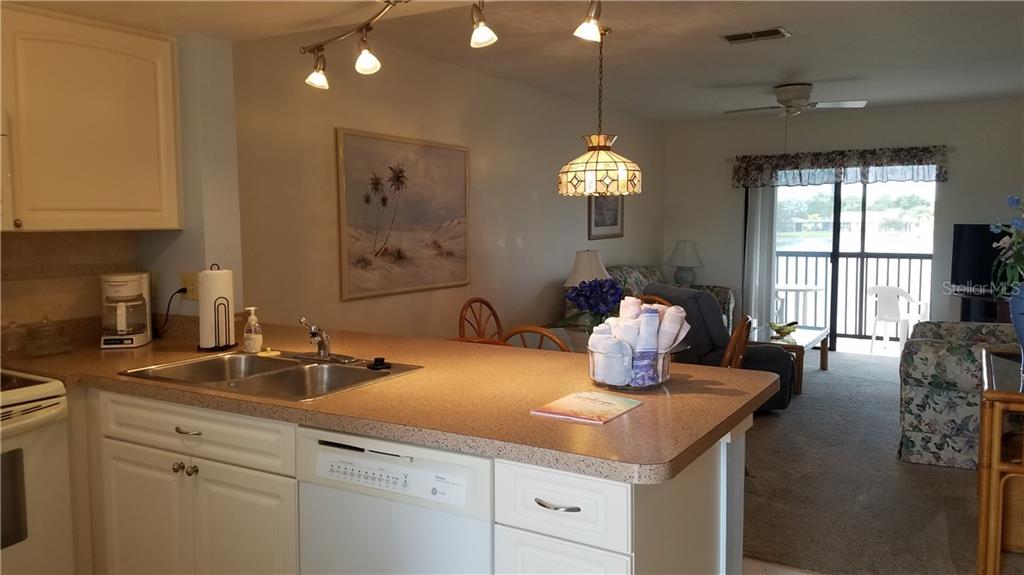 A kitchen with white cabinets and a breakfast bar overlooks a living room with balcony — 25275 RAMPART BOULEVARD UNIT 1608, PUNTA GORDA, FL