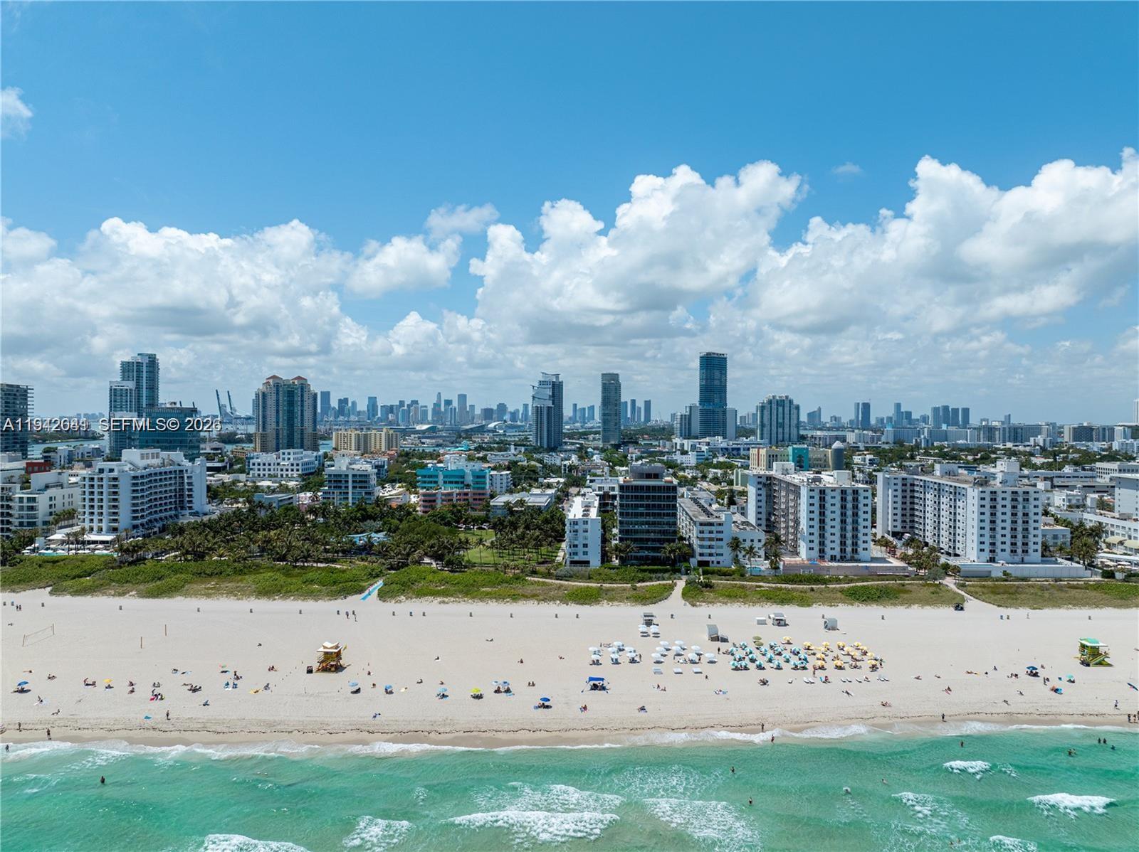 An aerial view shows a sandy beach with umbrellas and a city skyline in the background — 301 Ocean Dr # 302, Miami Beach FL 33139, MIAMI BEACH, FL