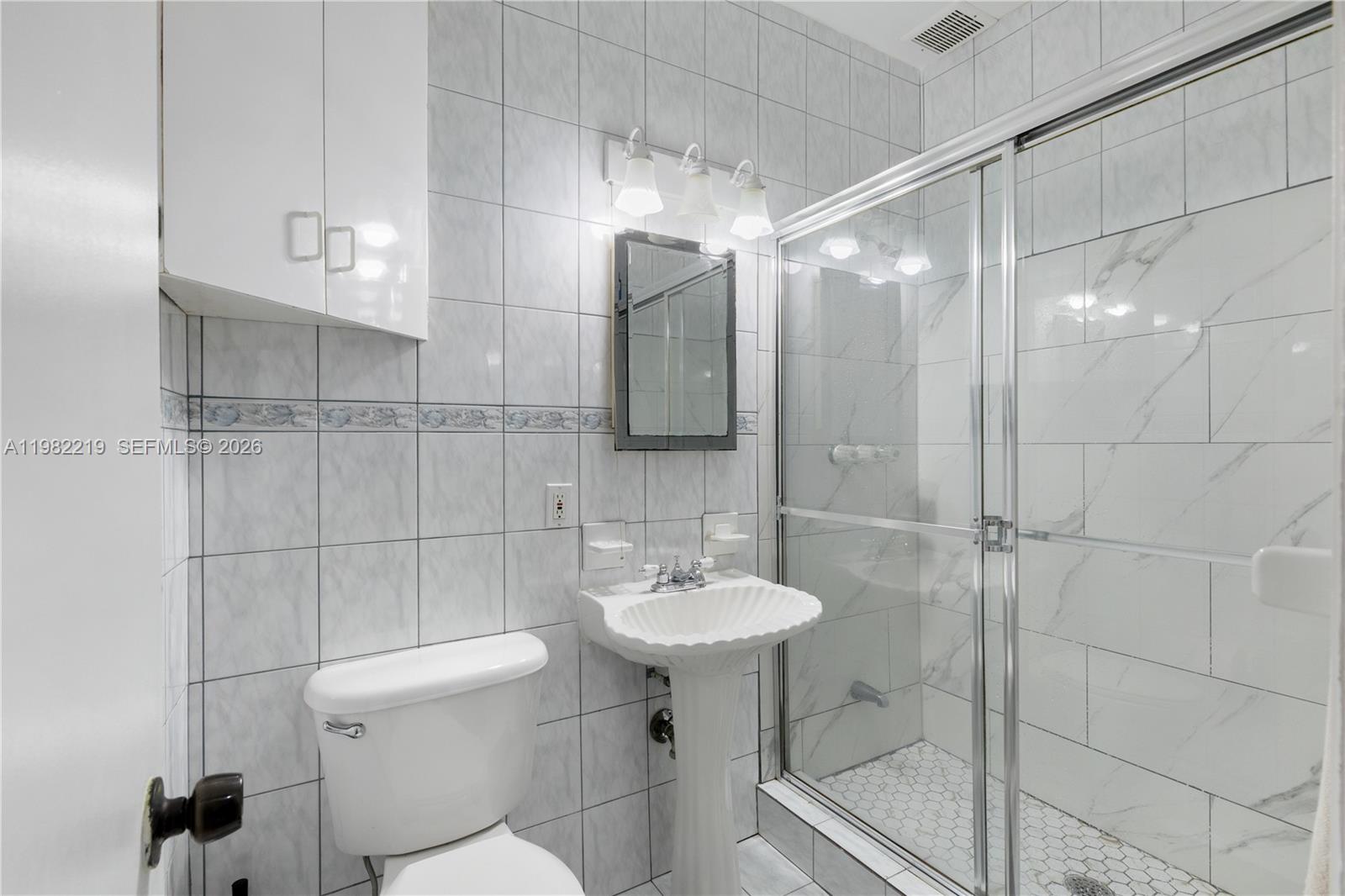 A bathroom features a pedestal sink, a toilet, and a glass-enclosed shower with tiled walls — 2445 Lake Pancoast Dr # J, Miami Beach FL 33140, MIAMI BEACH, FL