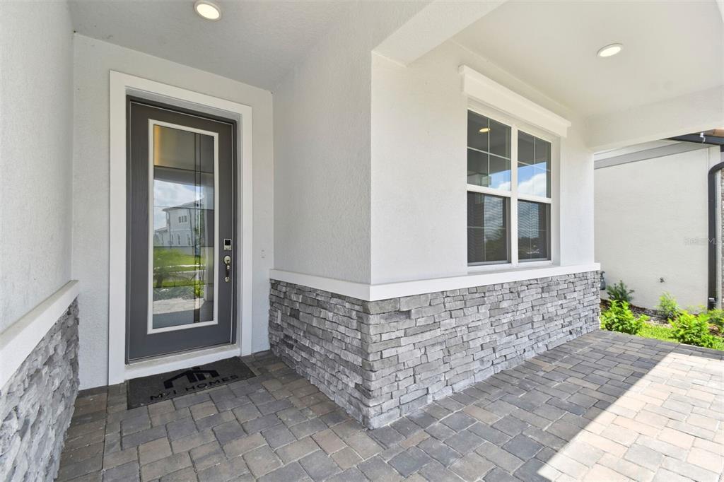A covered porch features a gray front door, stone veneer siding, and paver flooring — 1704 SUTTONSET TRAIL, ZEPHYRHILLS, FL